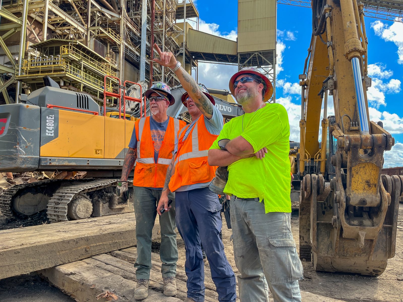 Three NADC team members in hard hats and safety vests reviewing work beside excavators at an industrial jobsite
