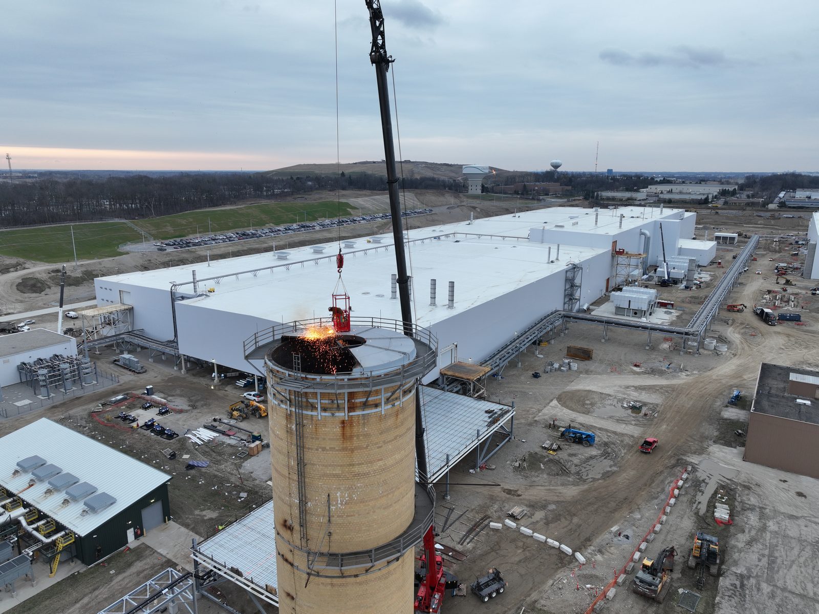Aerial view of crews torch-cutting the top of a brick stack beside a newly re-roofed automotive manufacturing plant