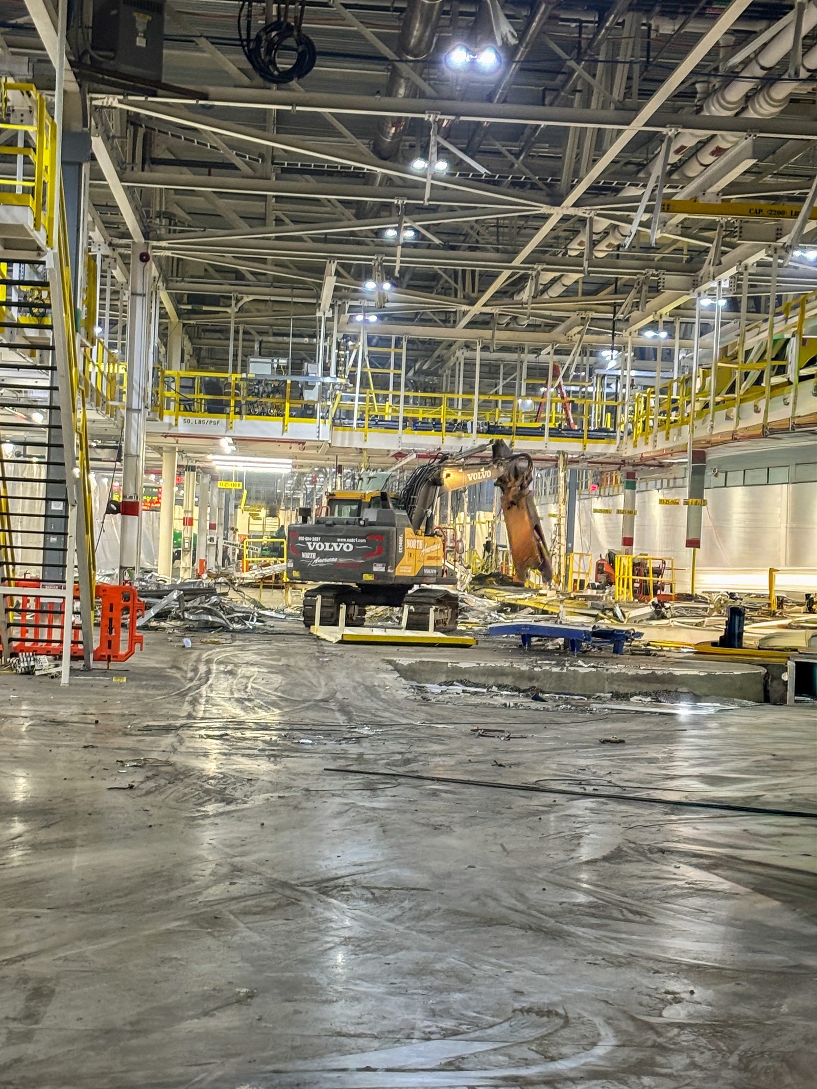 NADC excavator with shear attachment clearing production equipment inside an automotive plant beside elevated conveyor platforms