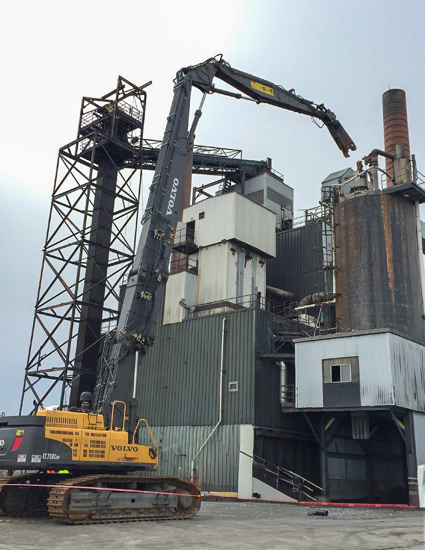 NADC high-reach excavator reaching over a process building beside a stack at an industrial plant