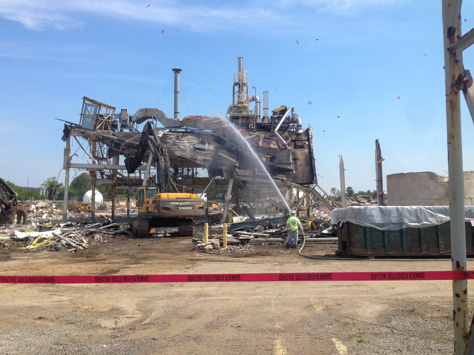 Excavator working through collapsed steel process structures behind hazard tape during chemical plant demolition