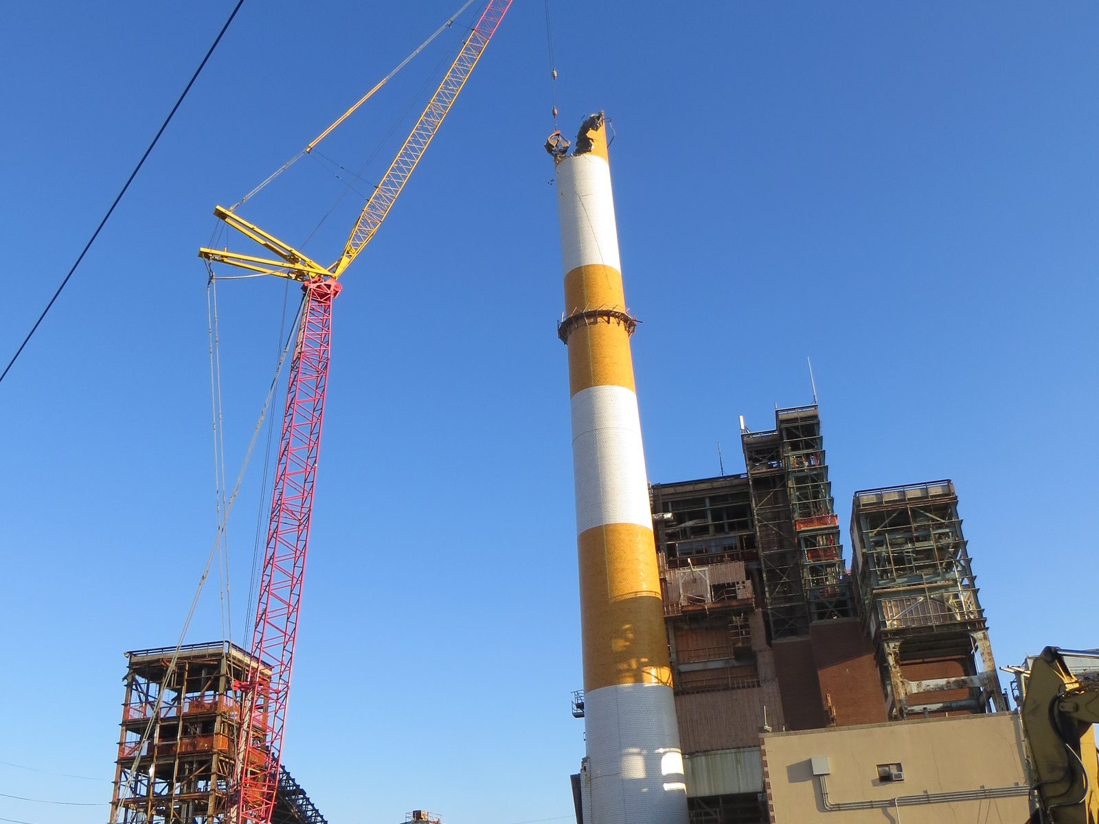 Lattice-boom crawler crane positioned beside a banded stack at a partially demolished power plant