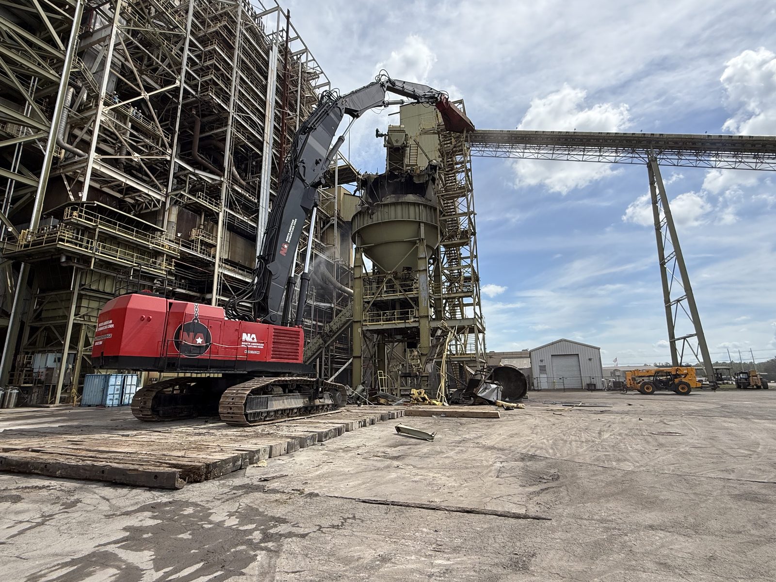 NADC high-reach excavator dismantling a steel process structure beside an elevated conveyor bridge