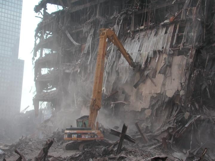 Long-reach excavator pulling down a heavily damaged multistory structure in thick dust