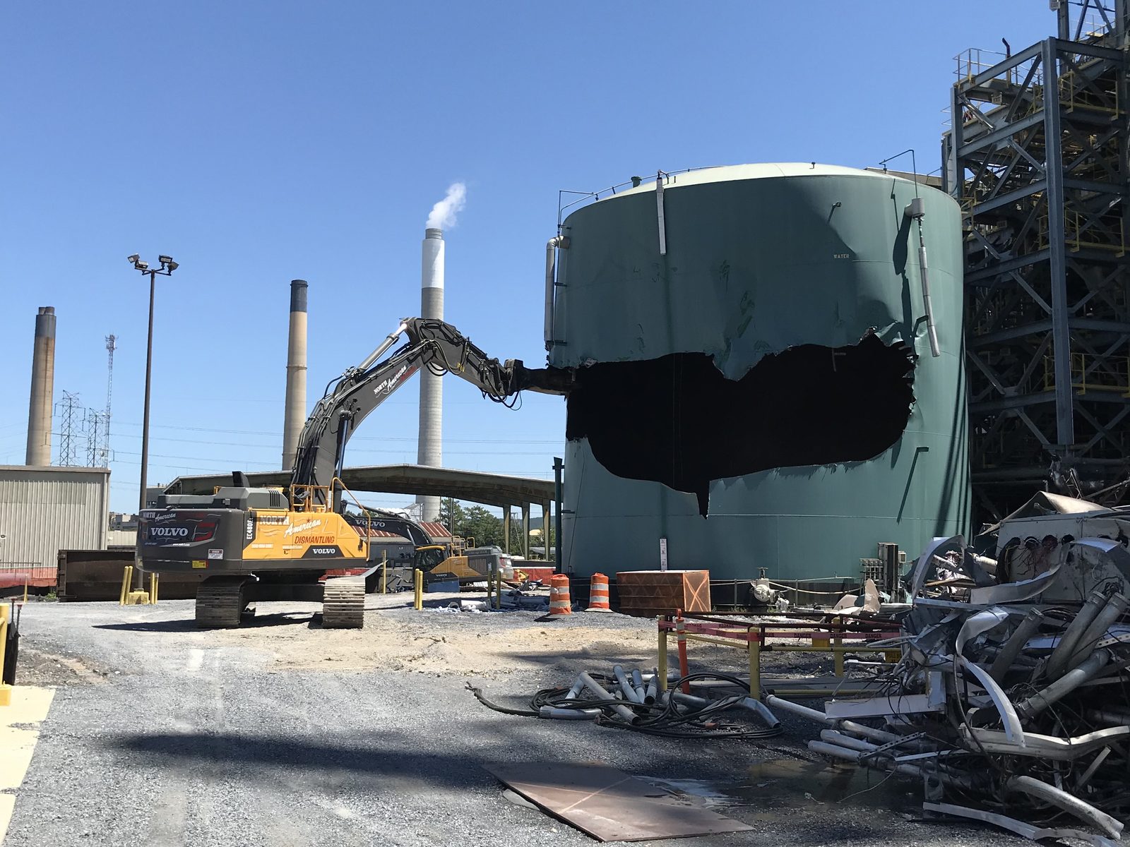 Excavator with shear cutting open a large green steel storage tank at an industrial plant